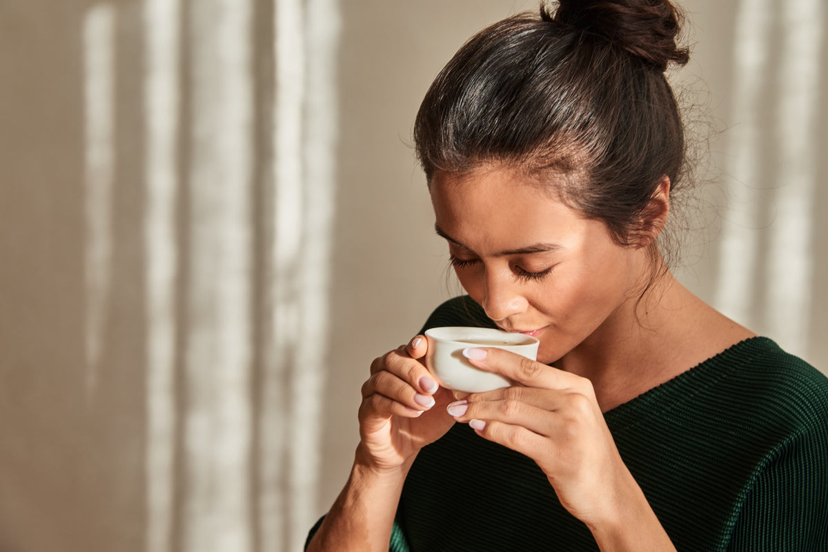 Person mit dunkelgrünem Oberteil hält eine kleine weiße Teetasse nah an den Mund; der Hintergrund zeigt vertikale Lichtmuster