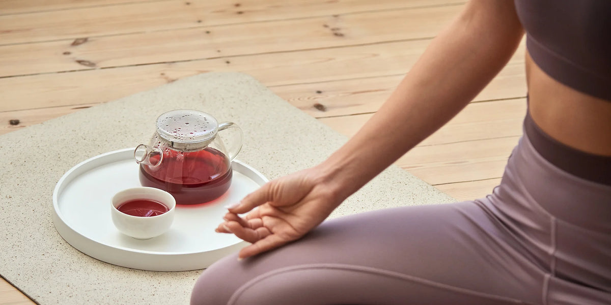Person im Meditationssitz auf Yogamatte, vor sich ein weißes Tablett mit Glasteekanne und Teetasse mit rotem Tee auf Holzfußboden