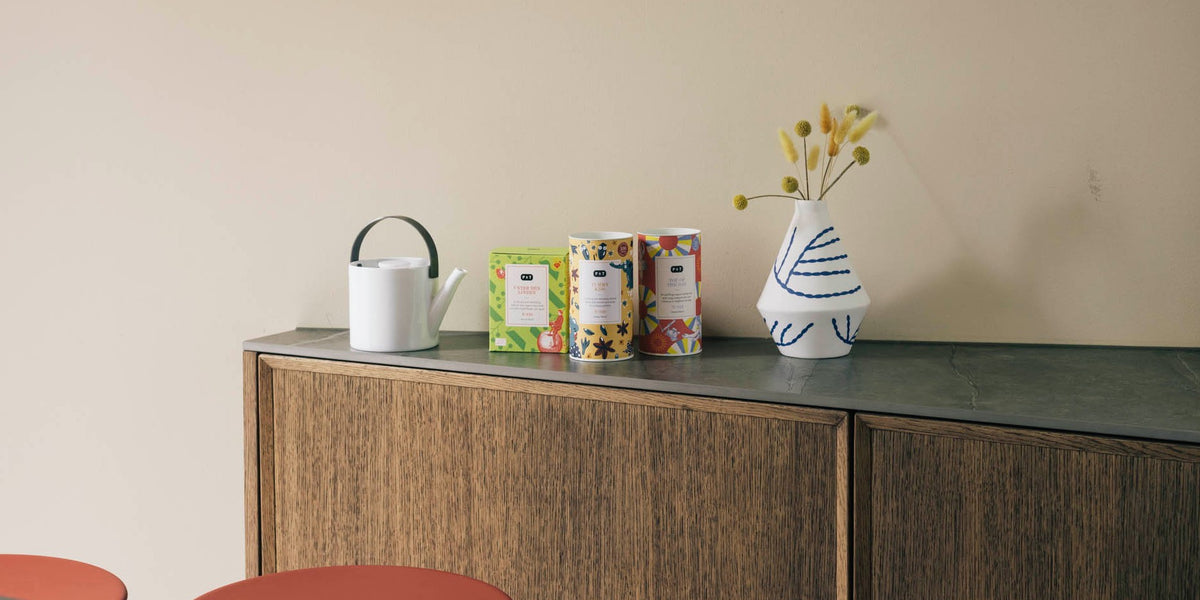 Holzschrank mit dunkler Oberfläche, darauf eine weiße Gießkanne, drei farbenfrohe Teeboxen mit unterschiedlichen Designs und eine weiß-blau gestreifte Vase mit gelben Blumen