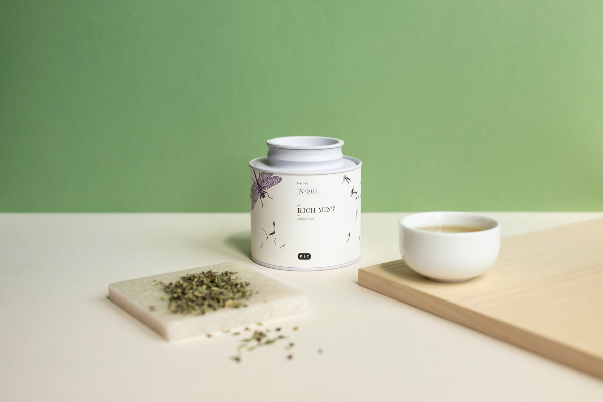 Weiße Teedose mit der Aufschrift ‚RICH MINT‘ und Illustrationen von Blättern und Schmetterlingen, daneben eine kleine weiße Schale mit Tee und eine quadratische Marmorplatte mit getrockneten Minzblättern vor grünem Hintergrund