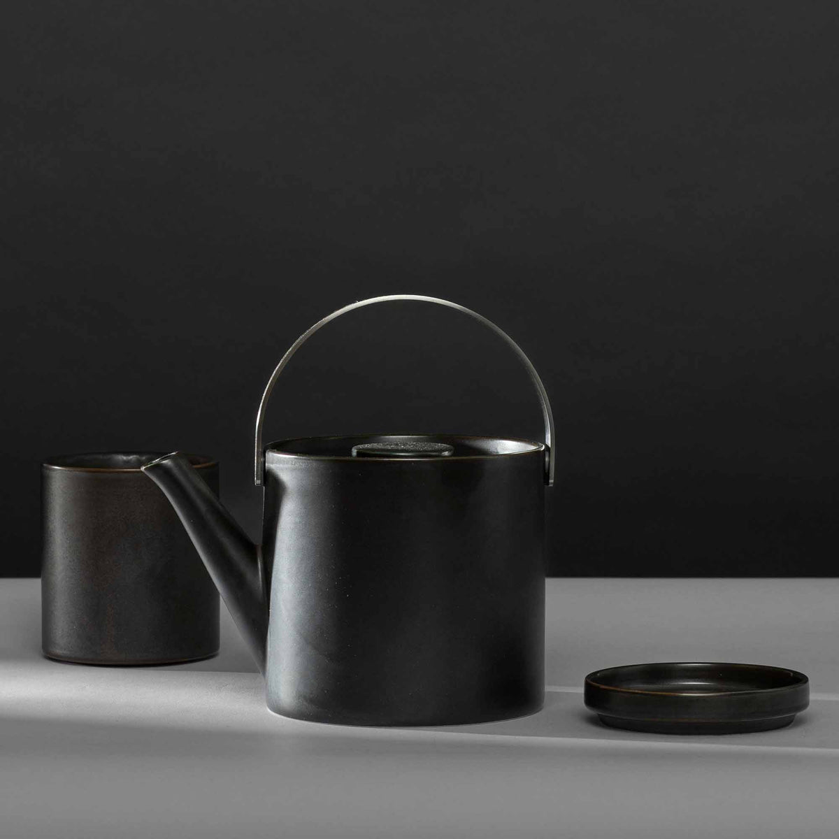Matte schwarze Teekanne mit gebogenem Griff, zylindrischer Becher und flacher Untersetzer minimalistisch arrangiert.