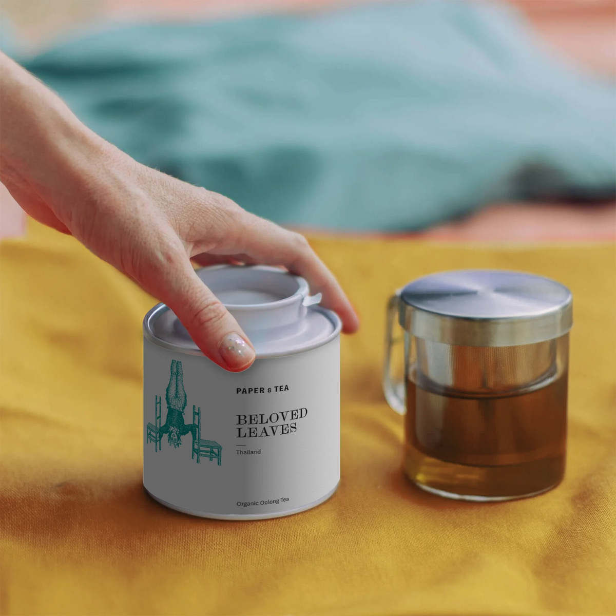 Eine Hand berührt den Deckel einer weißen Dose mit Bio-Oolong-Tee und einer Glastasse mit Teesieb.
