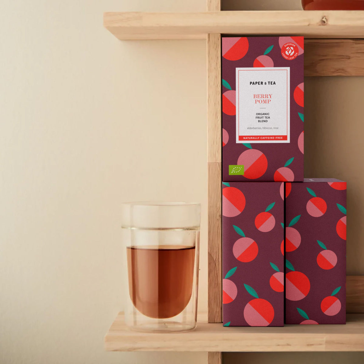 Drei stapelbare Beutel Paper & Tea Berry Pomp Früchtetee neben Glas mit rotem Tee auf Holzregal.