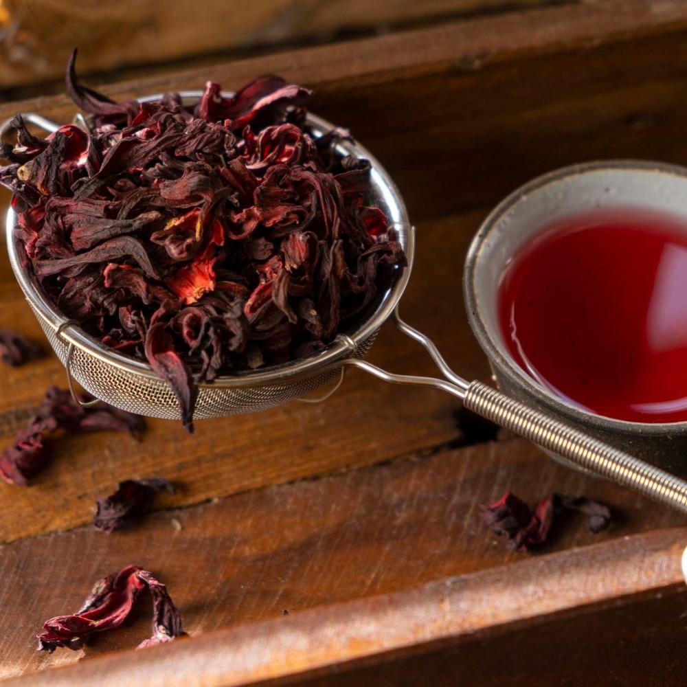 Metall-Teesieb mit getrockneten dunkelroten Hibiskusblüten über einem Holztablett mit einer Steintasse voller leuchtend rotem Hibiskustee.