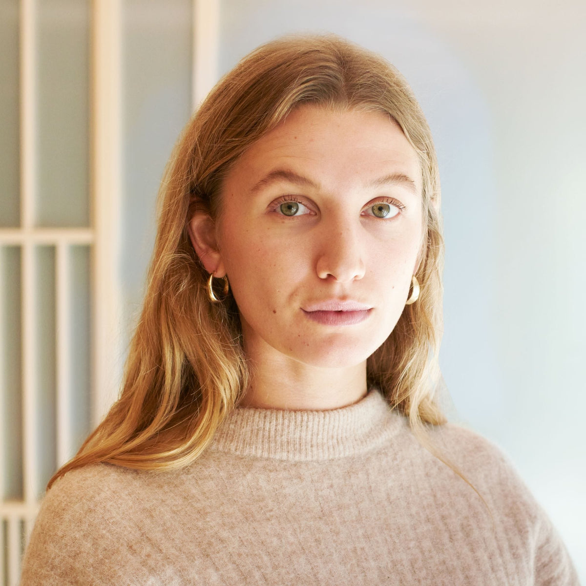 Junge Frau mit langen blonden Haaren und grünen Augen im beige gerippten Pullover mit goldenen Creolen.