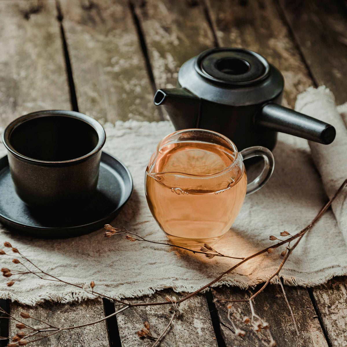 Rustikales Teeset mit Glas-Krug, schwarzer Keramikkanne, leerer Tasse auf Leinentuch mit getrocknetem Zweig.