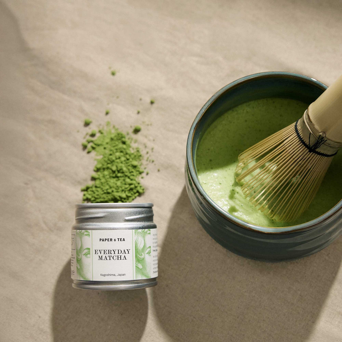 Metallbehälter mit Matcha-Pulver, Keramikschale mit Matcha-Tee und Bambusbesen auf beigem Stoff.