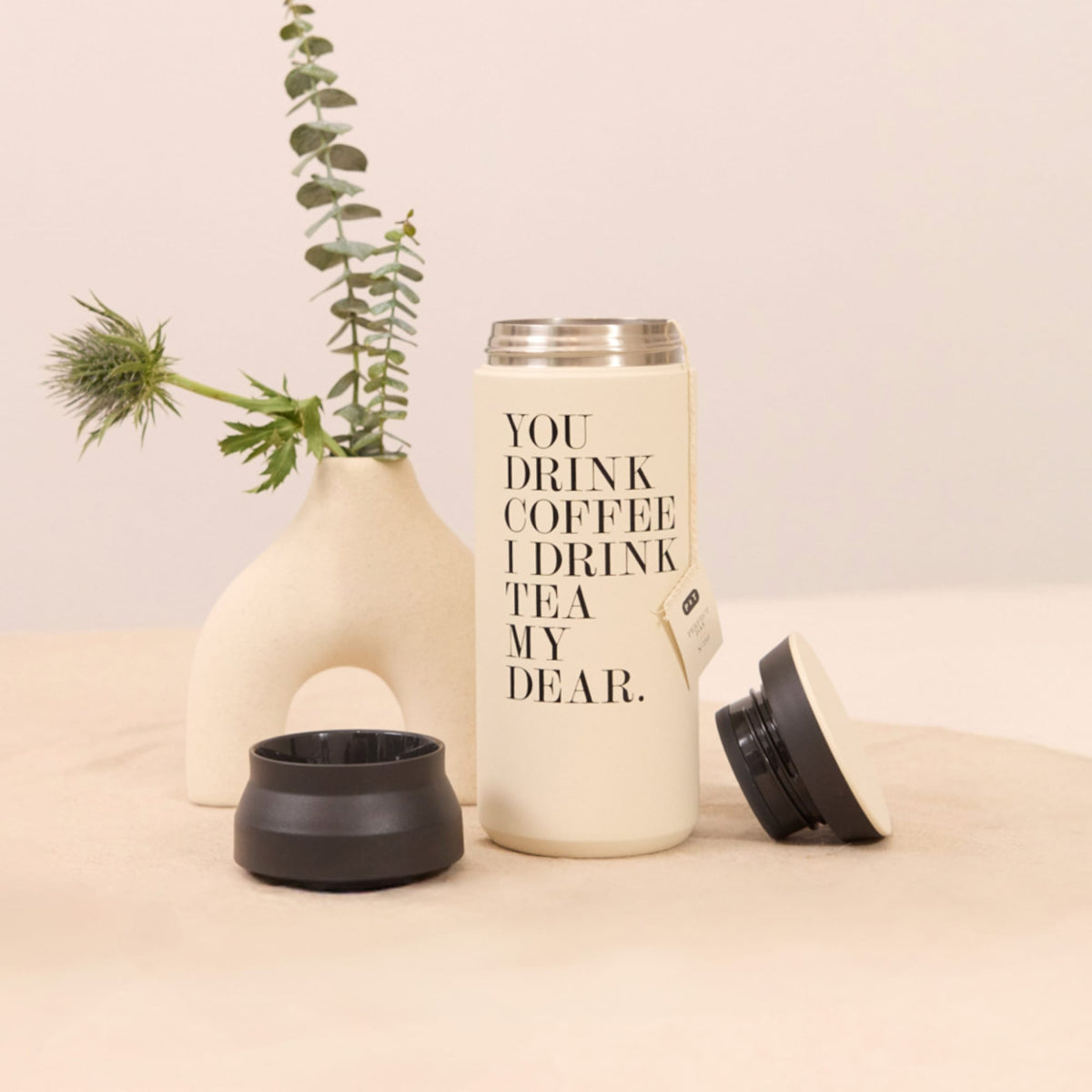 Beiger Trinkbehälter mit Aufschrift 'YOU DRINK COFFEE I DRINK TEA MY DEAR', zwei Deckel, Keramikvase mit Eukalyptus.