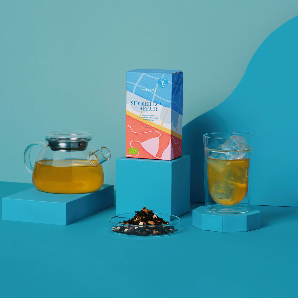 Bio-Eistee Summer Love Affair mit Glaskanne, Eisglas und Schwarztee vor blauem Hintergrund