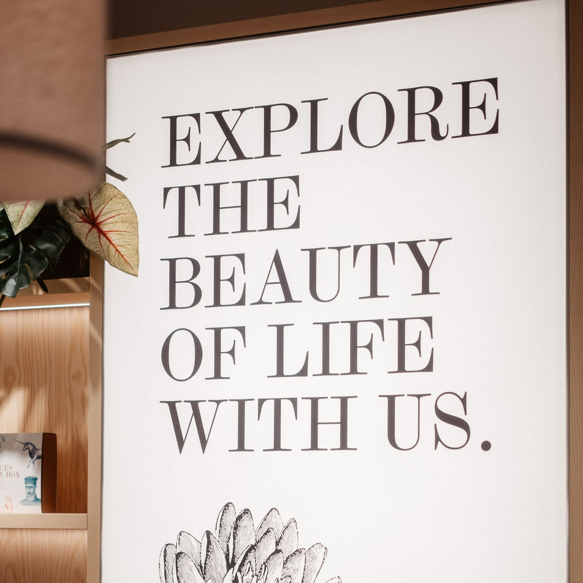 Großes weißes Poster mit ‚EXPLORE THE BEAUTY OF LIFE WITH US.‘ und Blumenillustration, neben Bücherregal.