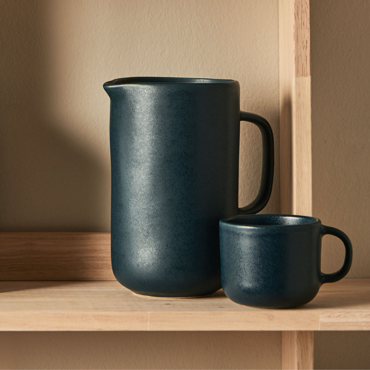 Dunkelblauer Keramikkrug mit Henkel und passendem Becher auf Holzregal vor beigefarbener Wand.