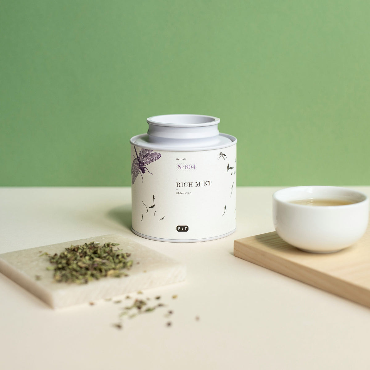 Weißer zylindrischer Kräutertee-Behälter mit botanischem Design, Rich Mint No 804, neben Keramiktasse mit hellem Tee.
