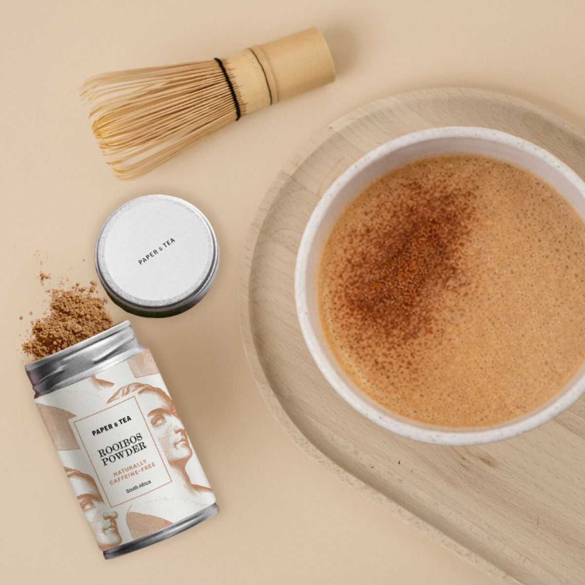 Metallbehälter mit losem Rooibos von P&T, Bambusbesen, Tasse mit schaumigem Rooibos-Tee auf Holztablett
