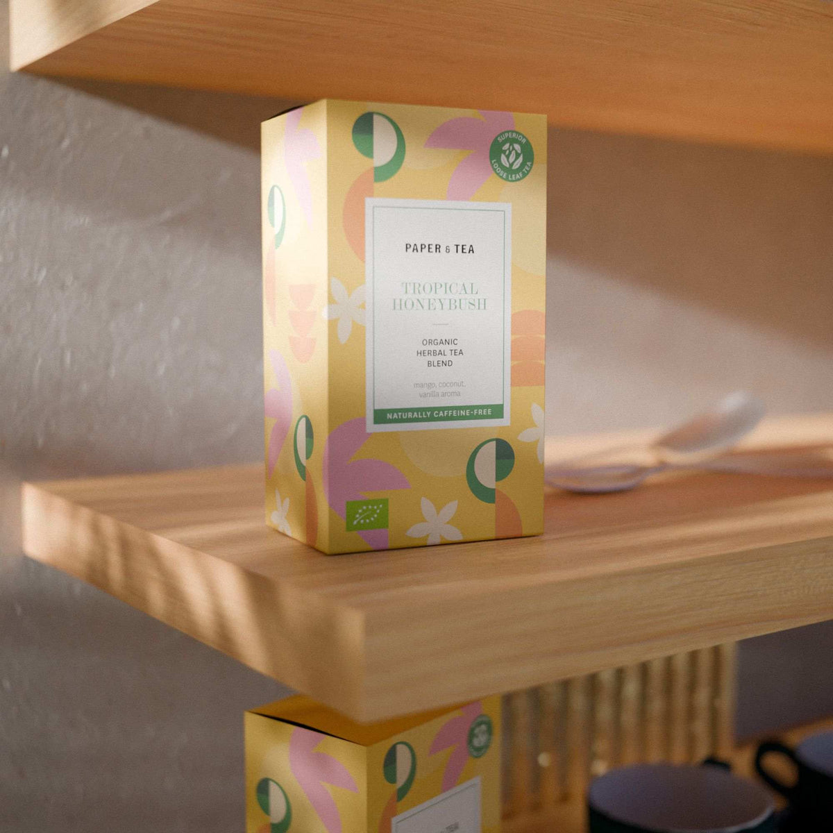Rechteckige Teebox für Tropical Honeybush Kräutertee von P&T, gelb-pink dekoriert, auf Holzregal.