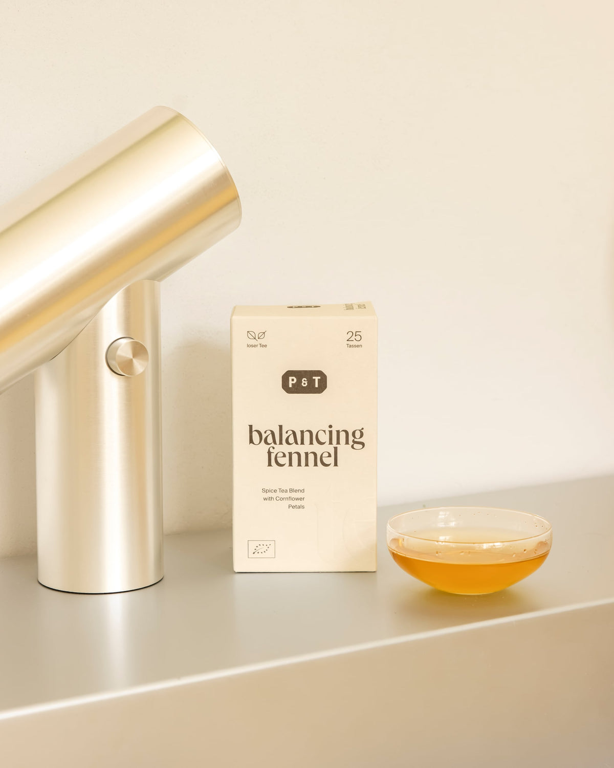 Beige P&T Teeverpackung mit Fenchel-Gewürztee, Kornblumenblüten, Teeaccessoire und bernsteinfarbener Tasse.