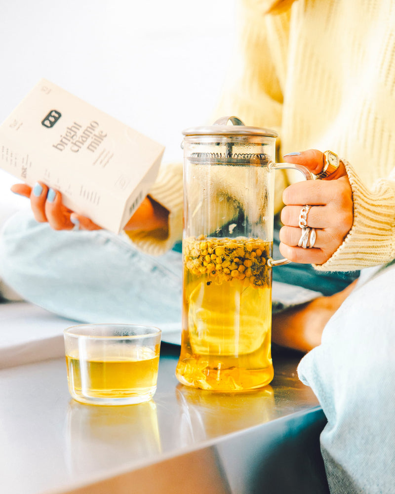 Person hält French Press mit leuchtend gelbem Kamillentee und Schachtel 'bright chamomile' in gemütlicher Umgebung.