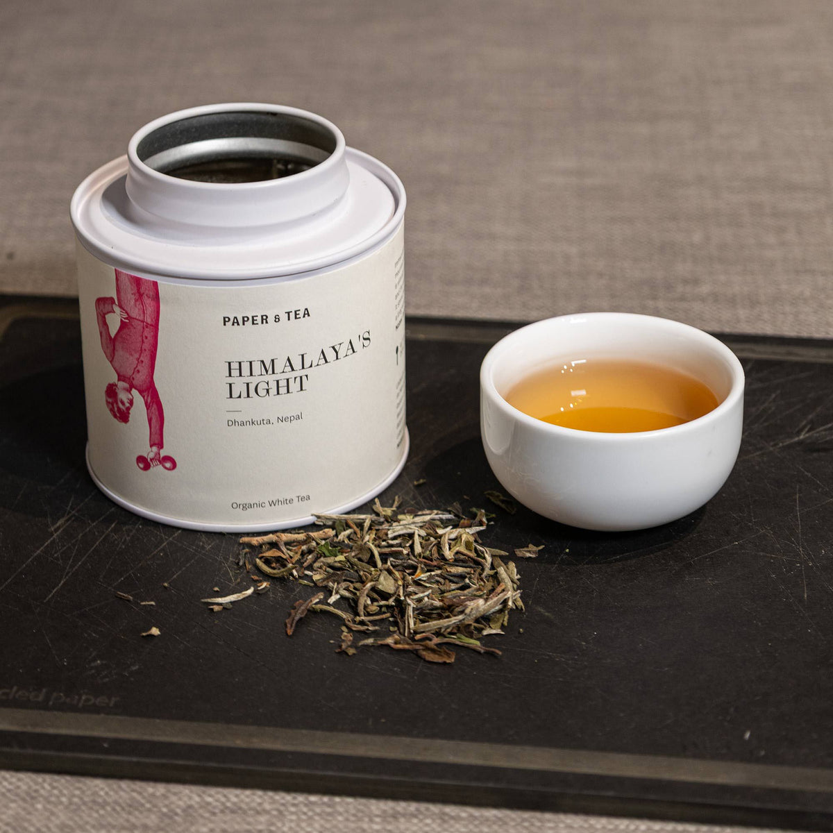 Weißer Teebehälter mit rotem Akrobat-Symbol, gefüllt mit Himalaya-Weißen Tee aus Nepal, neben Keramiktasse.