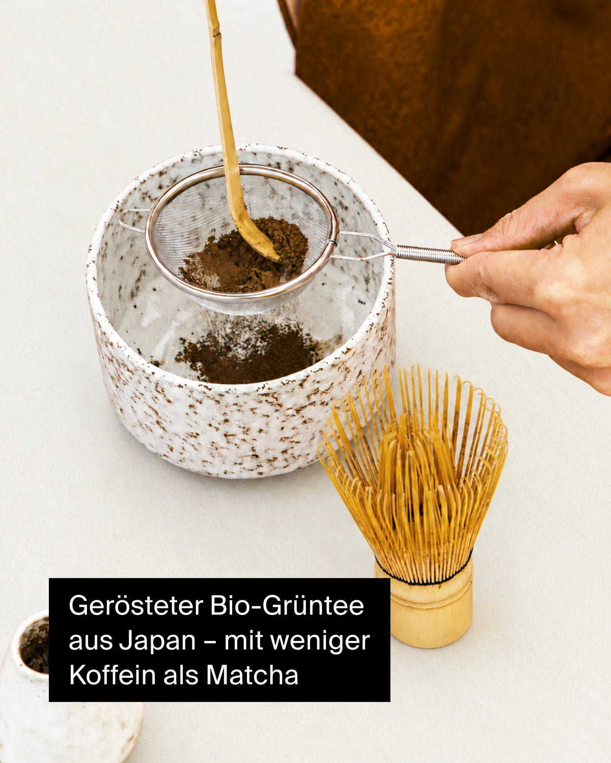 Weiß gesprenkelte Keramikschale mit geröstetem Bio-Grüntee, Metallsieb und Bambusbesen auf Beige.