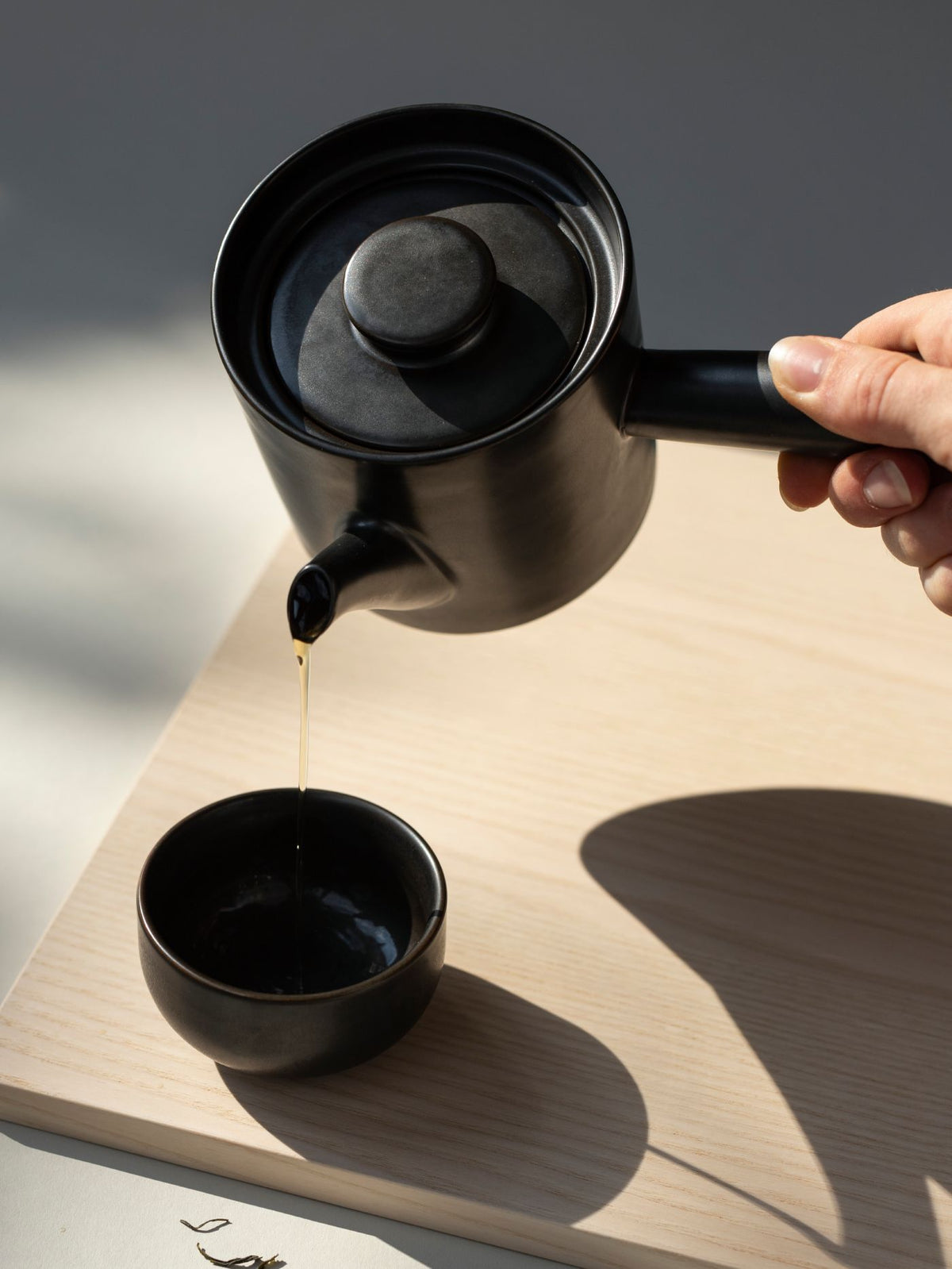 Schwarze Keramik-Teekanne gießt Tee in kleine schwarze Tasse auf hellem Holzbrett, Hand am Griff