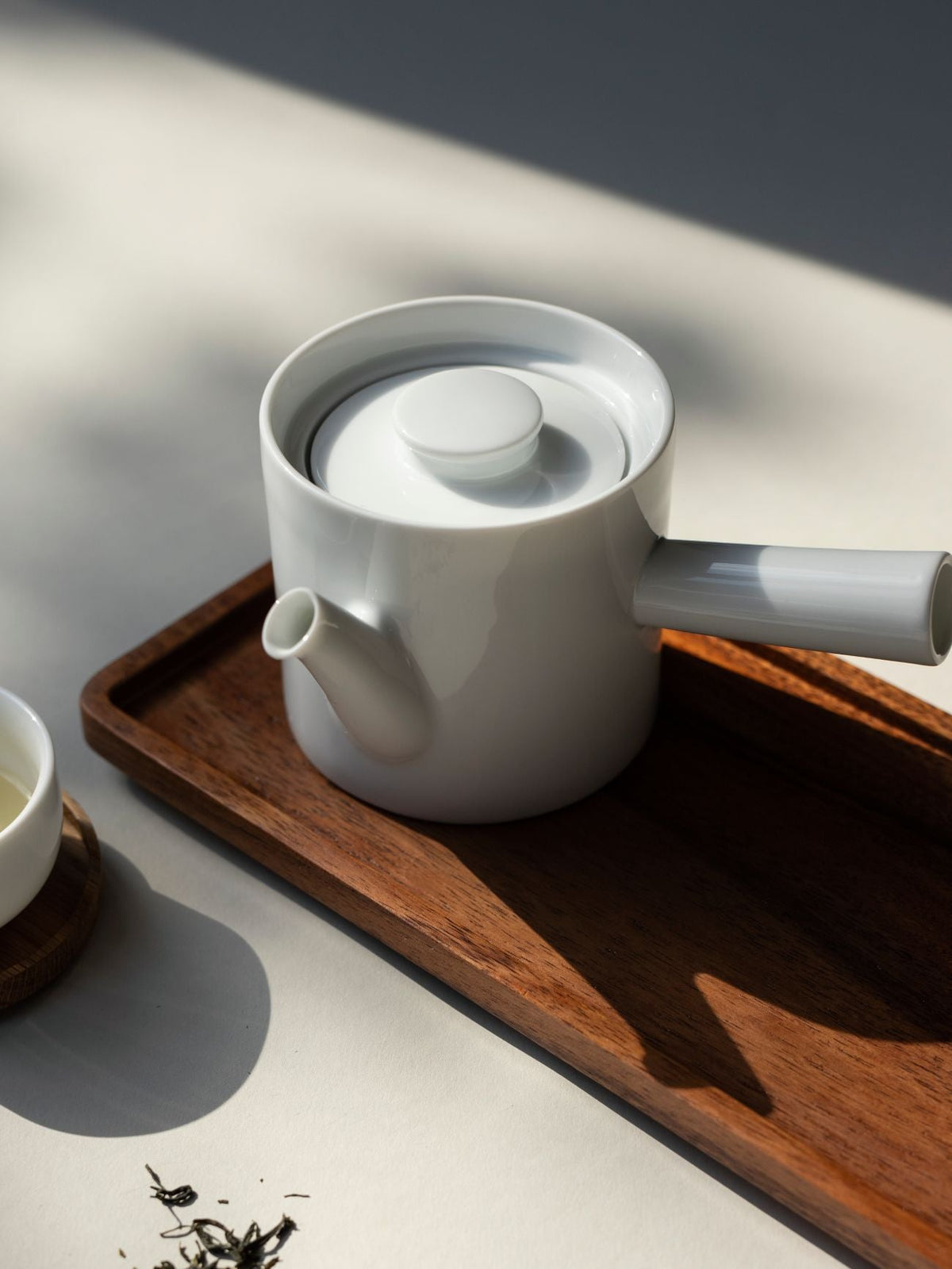 Weiße Keramik-Teekanne auf Holztablett mit Tasse und losen Teeblättern im natürlichen Licht.