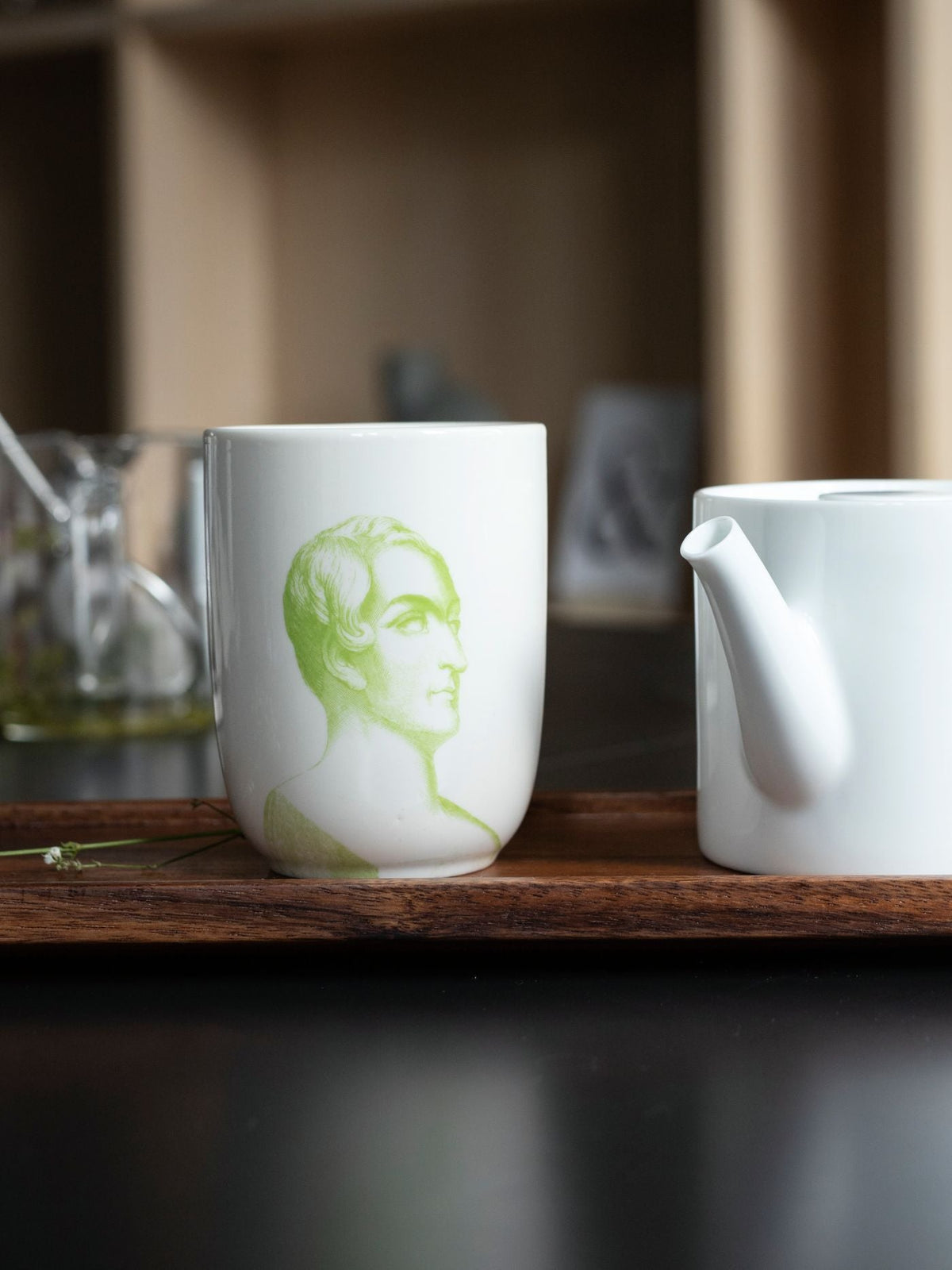 Weiße Keramiktasse mit grünem Männerprofil auf Holztablett neben weißem Teekännchen für minimalistisches Tee-Erlebnis.