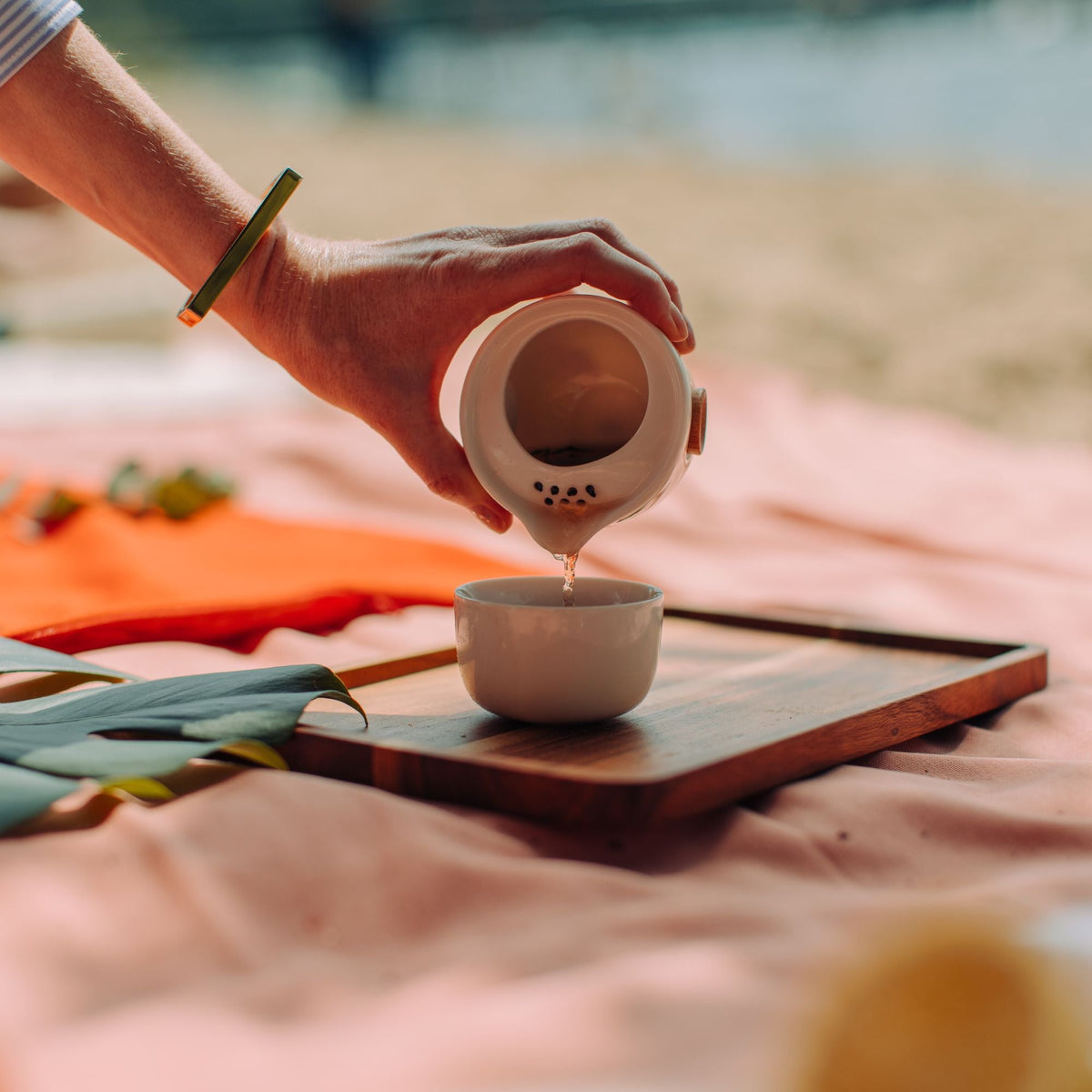 Hand mit goldenem Armband gießt Tee aus weißem Teekännchen in Tasse auf Strand-Holztablett.