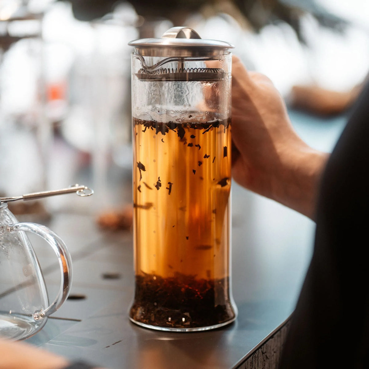 Glas French Press mit frisch gebrühtem Tee, gehalten von einer Hand auf einem Metalltisch.