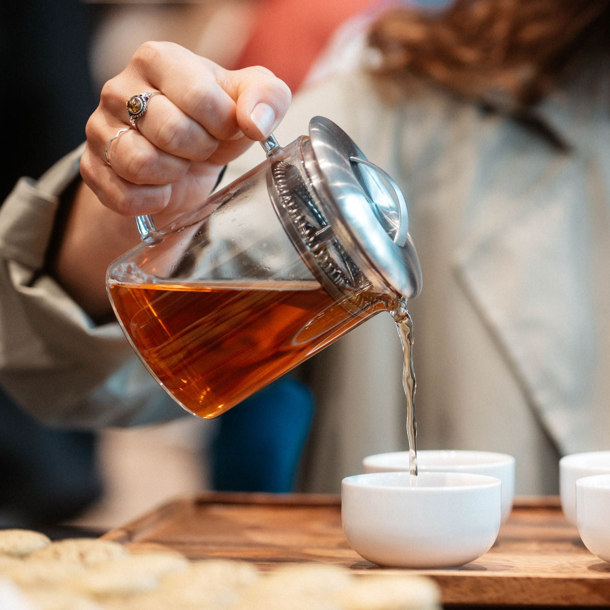 Eine Hand mit Ringen gießt warmen Tee aus einer Glaskanne in eine weiße Keramiktasse auf einem Holztablett.