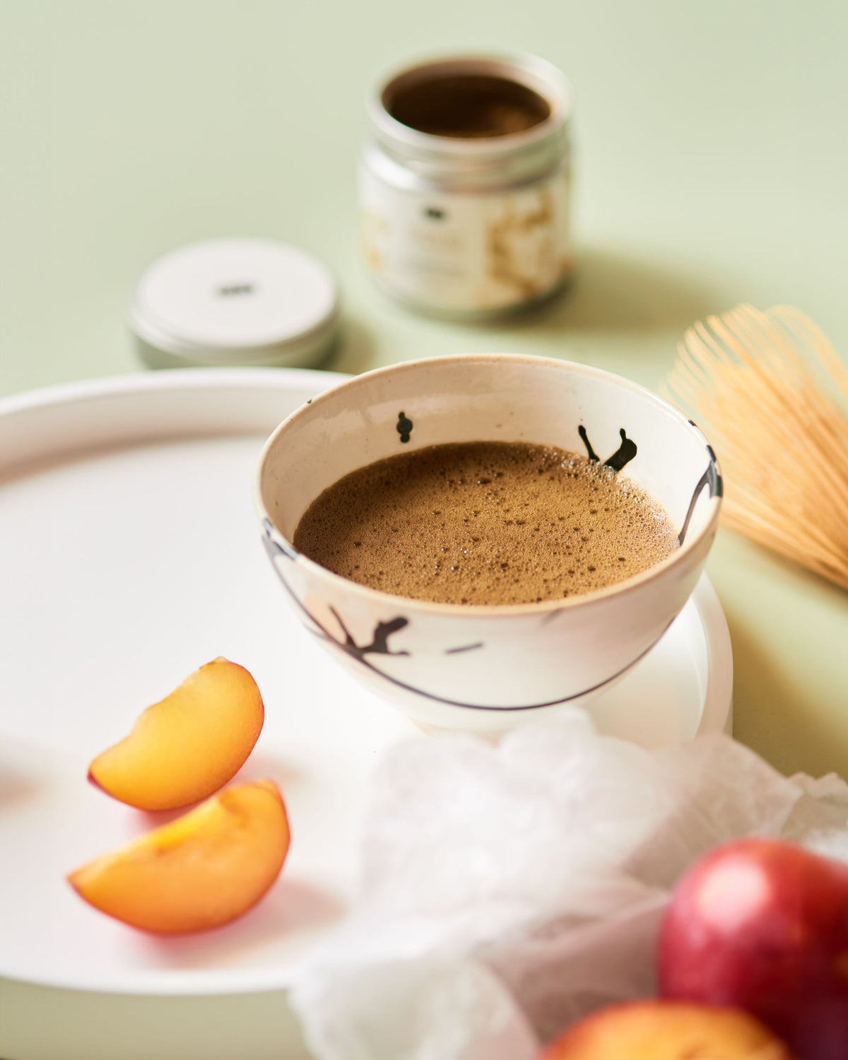 Rustikaler Keramikbecher mit floralem Muster und schaumigem Tee auf Tablett mit Zwetschgenscheiben und Teezubehör.