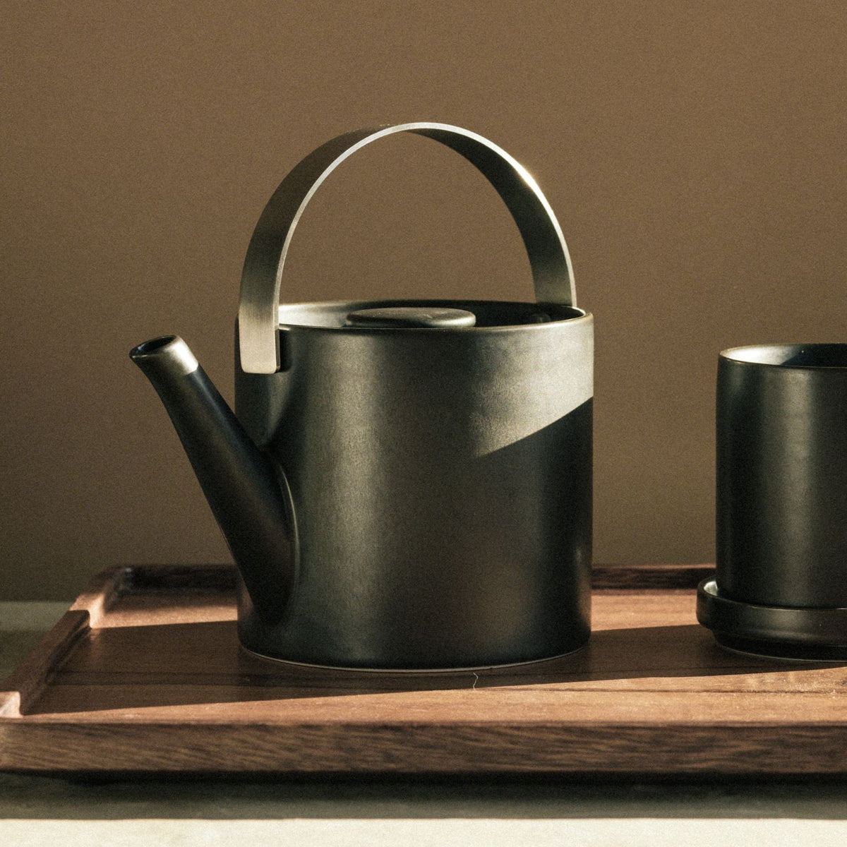 Mattschwarzes Teeset mit schlankem, zylindrischem Teekessel und passender Tasse auf Holztablett.