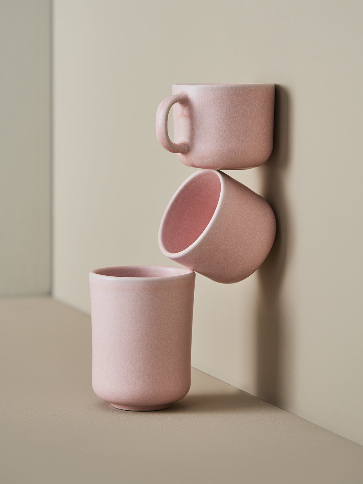 Drei matte rosa Keramiktassen, minimalistisch an beiger Wand arrangiert, eine mit Henkel oben, zwei henkelos unten.