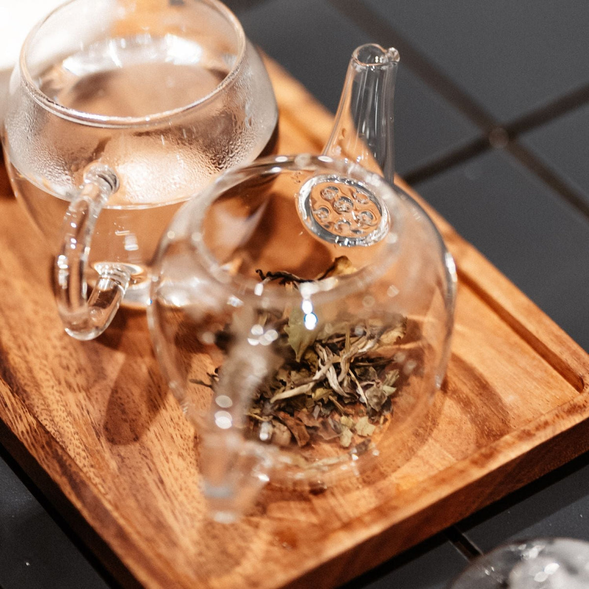 Glasteekanne mit losen Teeblättern und Glas heißem Wasser auf Holztablett auf kariertem Untergrund.