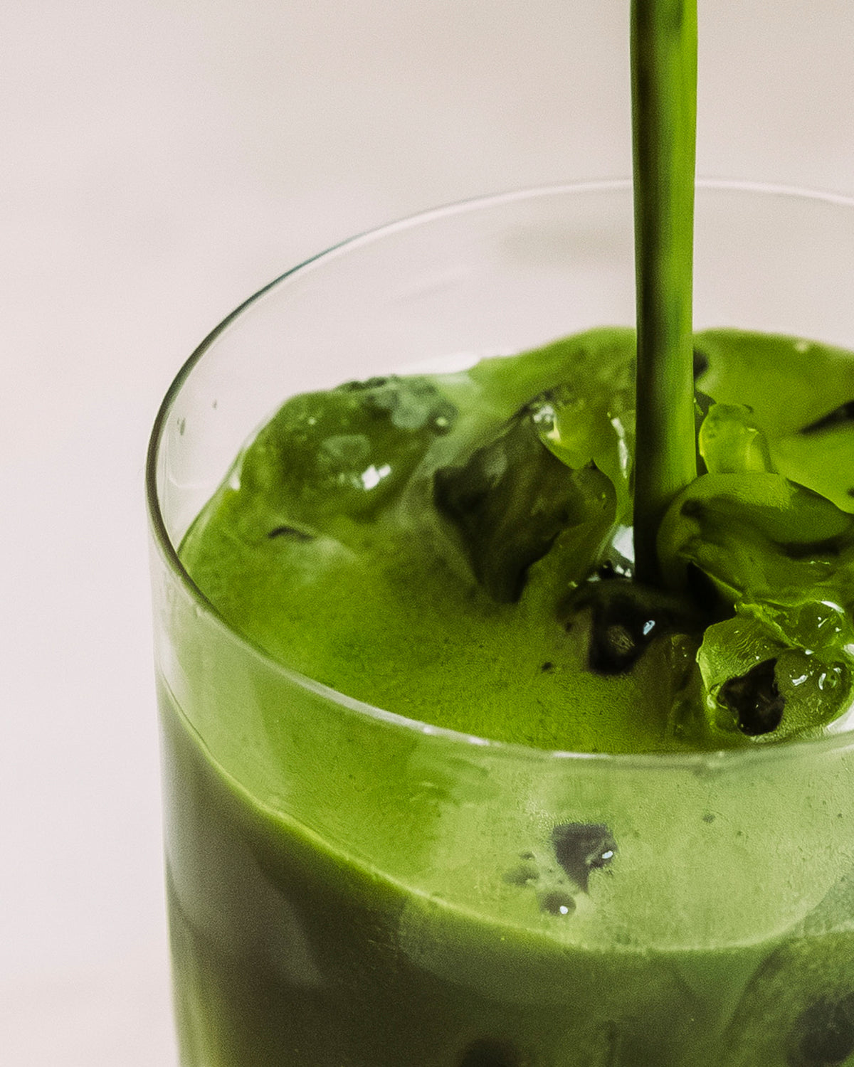 Ein Glas mit eisgekühltem grünem Getränk, in das frischer grüner Tee eingeschenkt wird, vor weißem Hintergrund.