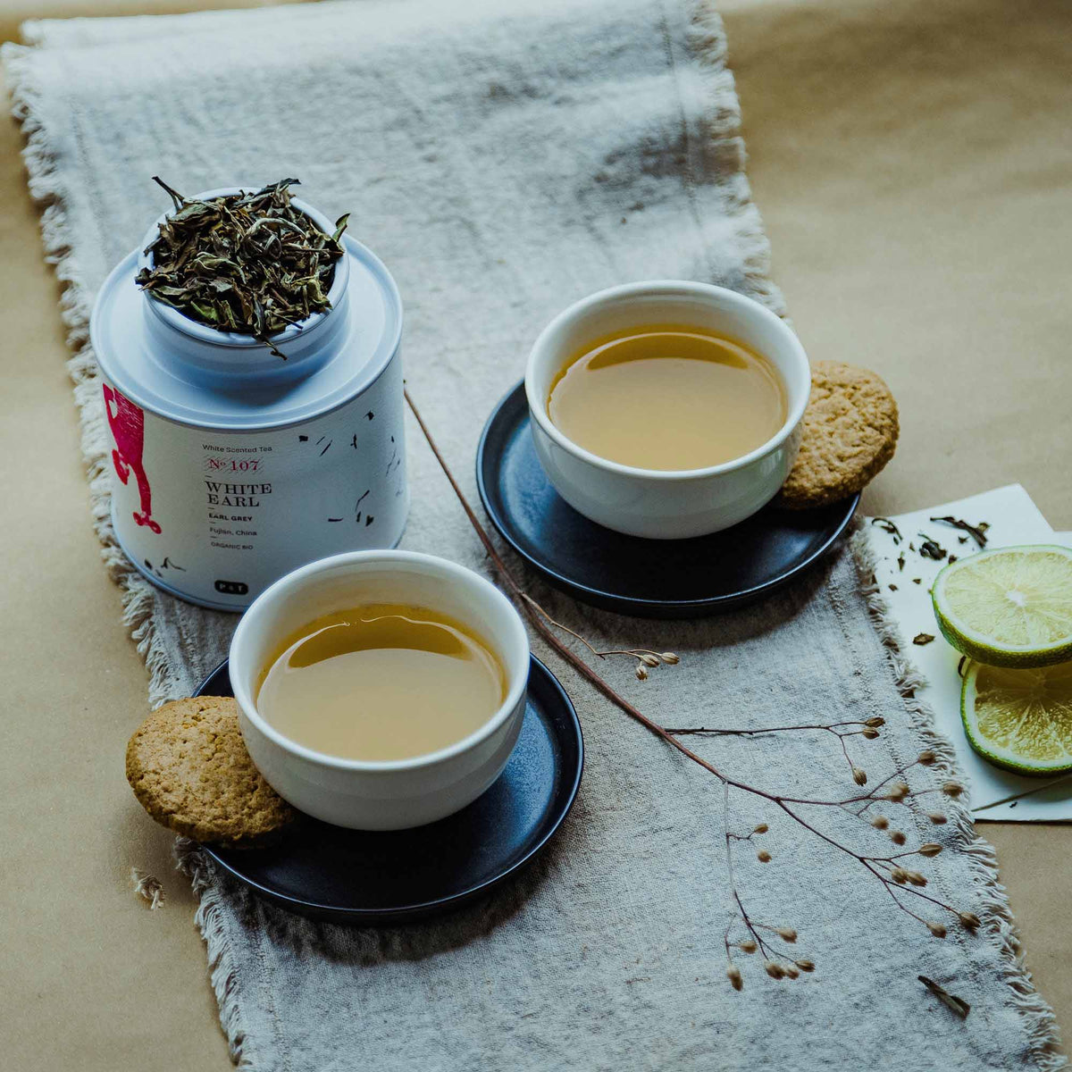 Weißes zylindrisches Teedöschen, zwei Tassen Earl Grey Tee, Kekse auf Untertassen, beiger Stoff, getrockneter Zweig, Limettenscheiben.