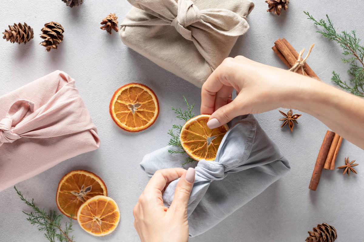 Drei Geschenkpakete in Rosa, Beige und Hellgrau, dekoriert mit getrockneten Orangen und Tannenzweigen