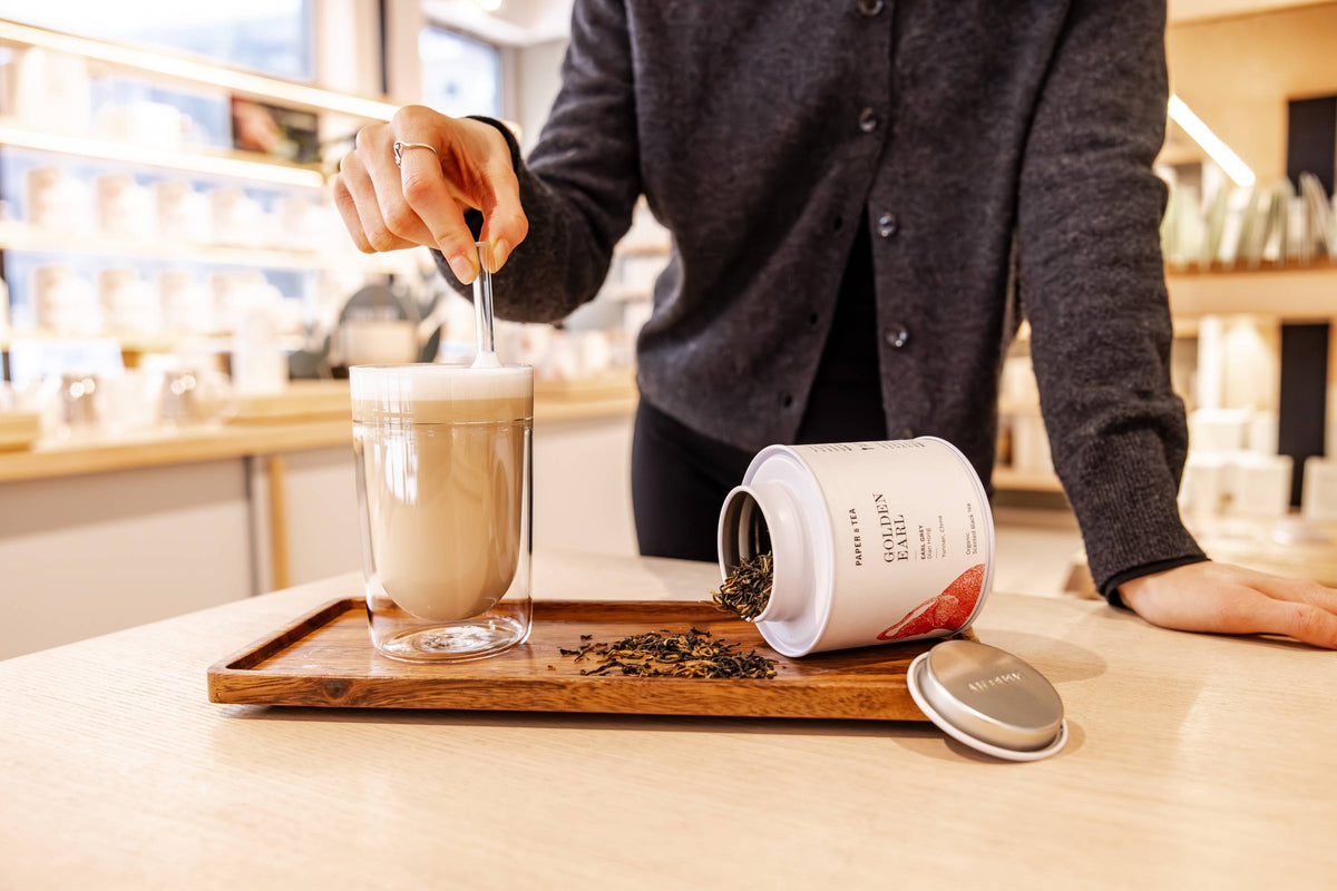 Person rührt in einem Glas schaumigem Earl Grey Getränk neben einer geöffneten Paper & Tea Teedose