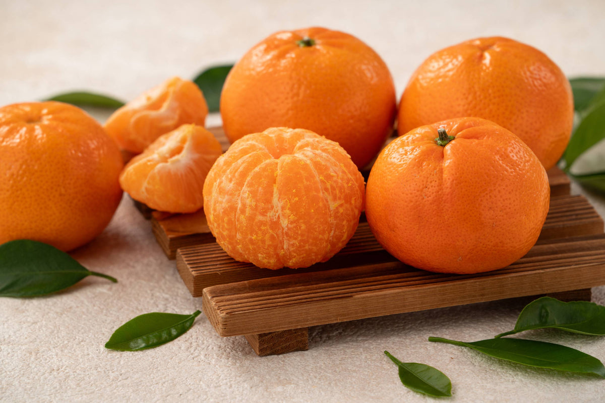 Fünf ganze Mandarinen und eine geschälte Frucht, umgeben von Zitrusblättern
