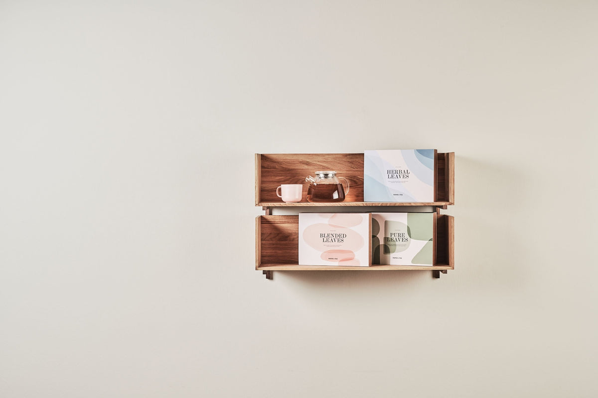 Wandregal aus Holz mit zwei Ebenen, darauf eine Glasteekanne, eine Teetasse und drei Teeboxen vor einer hellen Wand