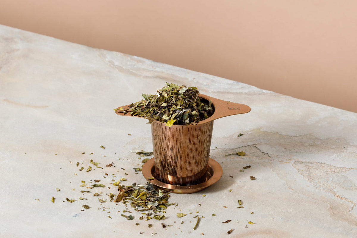 Kupferfarbener Tee-Infuser mit losen Teeblättern auf einer hellen Marmoroberfläche; einige Teeblätter sind rund um den Infuser verstreut