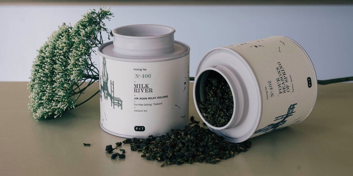 Zwei zylindrische Teedosen, eine stehend mit der Aufschrift ‚Oolong Tea No. 104 Milk River – Jin Xuan Oolong Tea Taiwan‘, die andere liegend mit geöffnetem Deckel und herausfallenden Teeblättern, daneben ein Zweig mit weißen Blüten