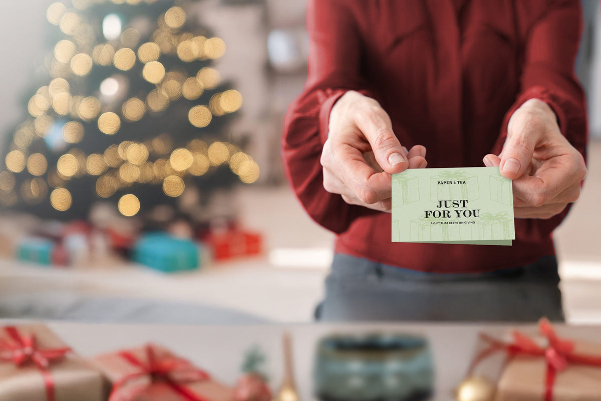 Hand hält eine grüne Geschenkkarte mit der Aufschrift ‚A GIFT FOR YOU‘ und ‚PAPER + TEA‘ vor gelbem Hintergrund
