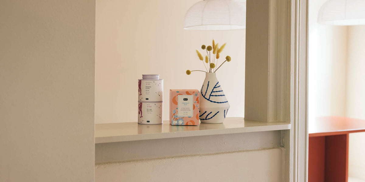Zwei gestapelte weiße Teedosen, eine orangefarbene quadratische Teeverpackung und eine Vase mit gelben Kugelblumen vor beiger Wand