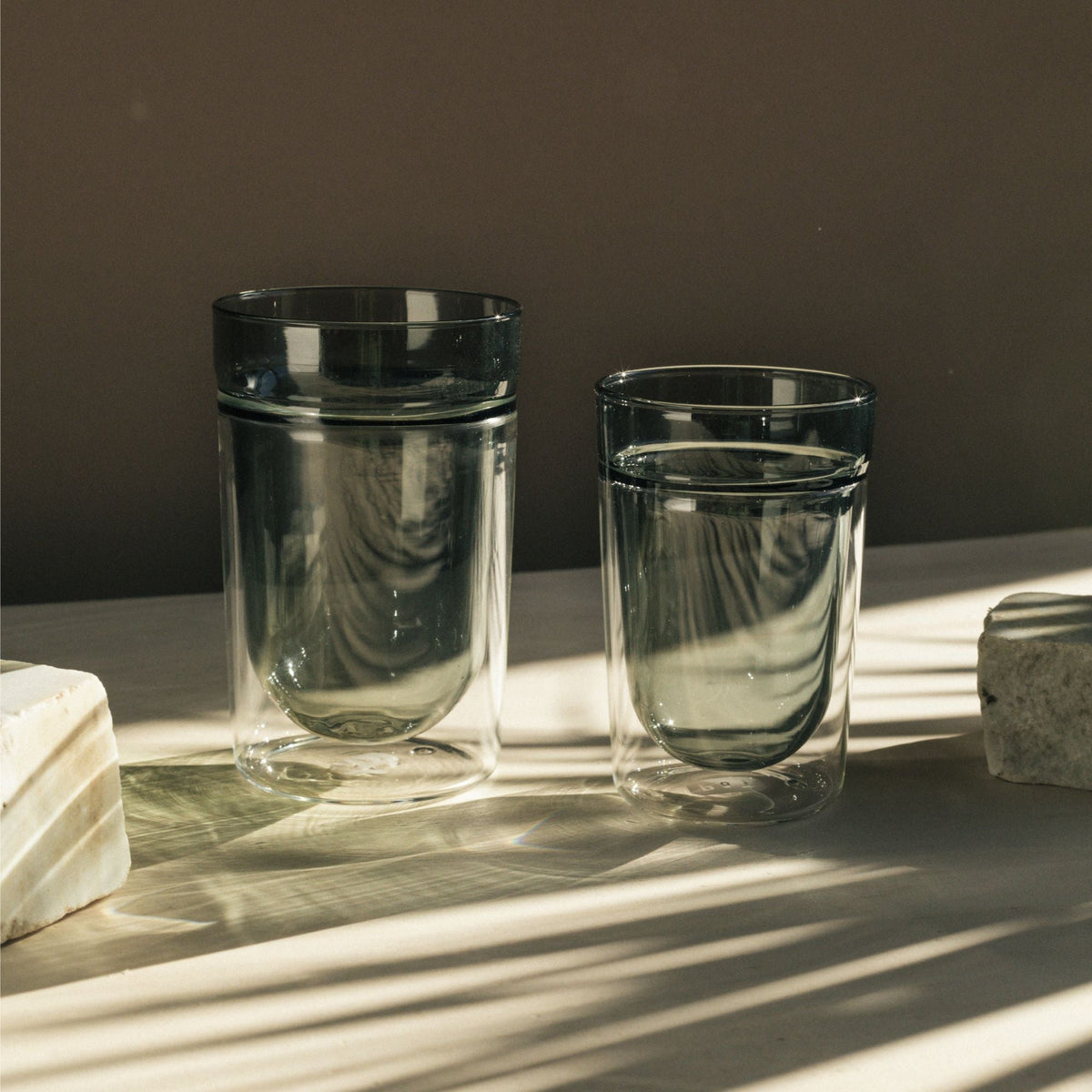 Zwei transparente Doppelwandgläser mit Wasser auf neutraler Oberfläche, umgeben von rechteckigen Steinblöcken.