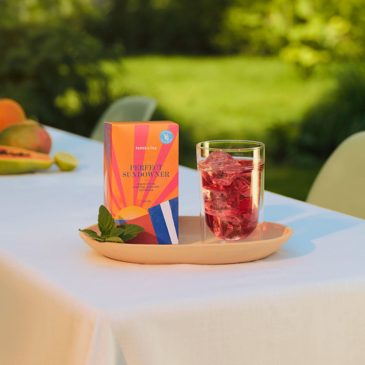 Orange Schachtel mit PAPER & TEA PERFECT SUNDOWNER Bio-Eistee, neben Glas mit rotem Eistee und Minze auf weißem Tischtuch.