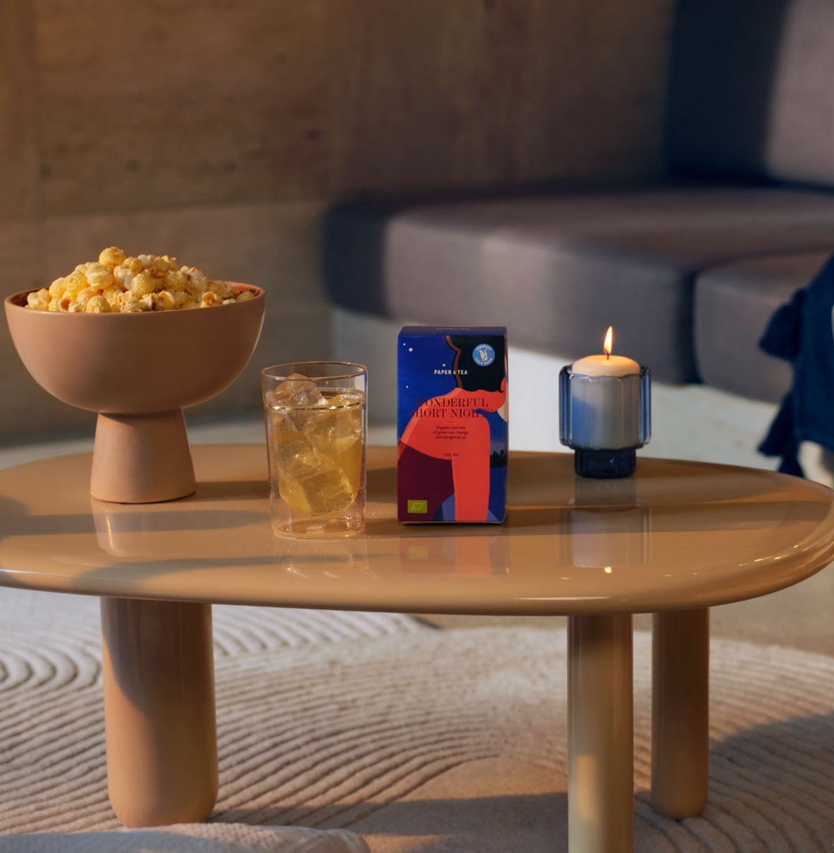 Beiger runder Couchtisch mit "PAPER TEA WONDERFUL HORT NIGHT" Tee, Popcorn, Eistee und Kerze im Wohnzimmer.