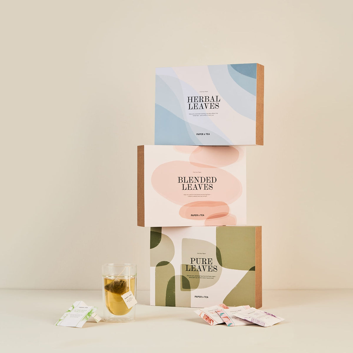 Drei pastellfarbene Paper & Tea Teeboxen, ein Glas Tee und verstreute Teebeutel auf neutralem Hintergrund.