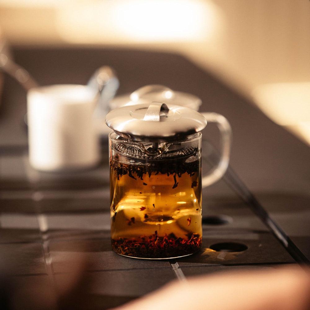 Klarer Glas-Teekessel mit Metallsieb und Teeblättern auf dunklem Tisch, unscharfe weiße Tasse im Hintergrund.