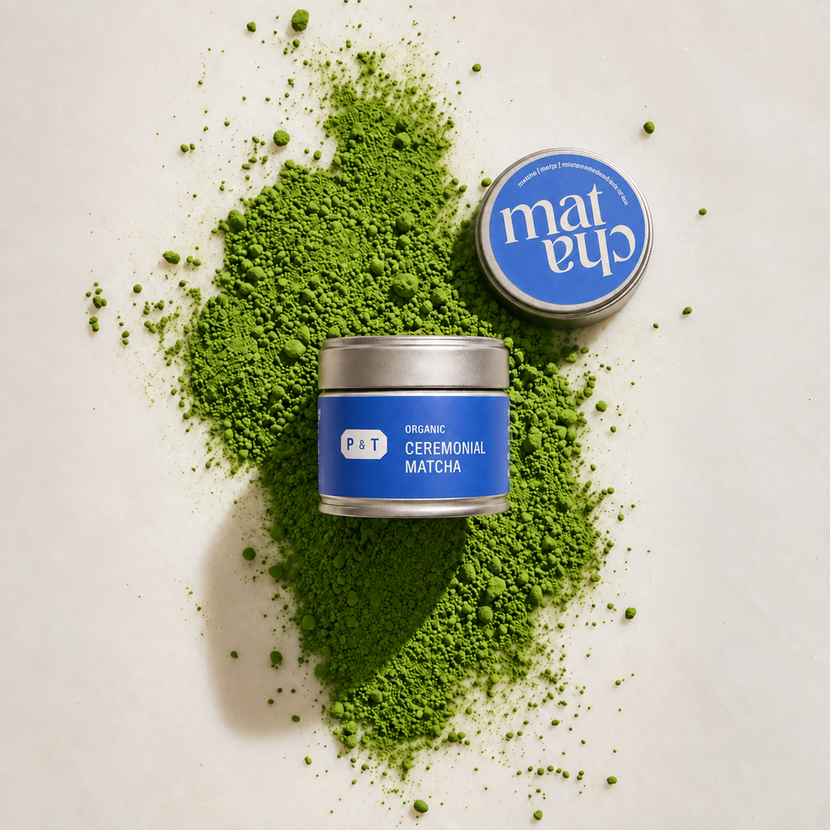 Zylindrische Metalldose mit blauem Etikett 'P&T ORGANIC CEREMONIAL MATCHA' auf hellem Untergrund, grünes Matcha-Pulver, verschlossener Deckel daneben