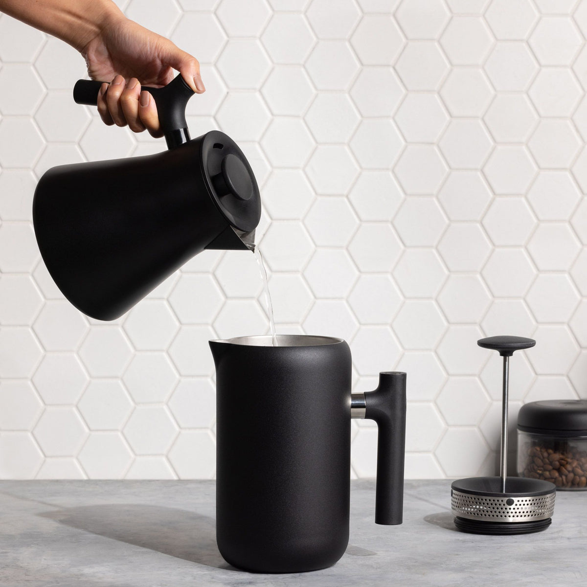 Mattschwarze French Press auf hellem Küchentresen, Wasser wird hineingegossen, minimalistisches Kaffeezubehör.