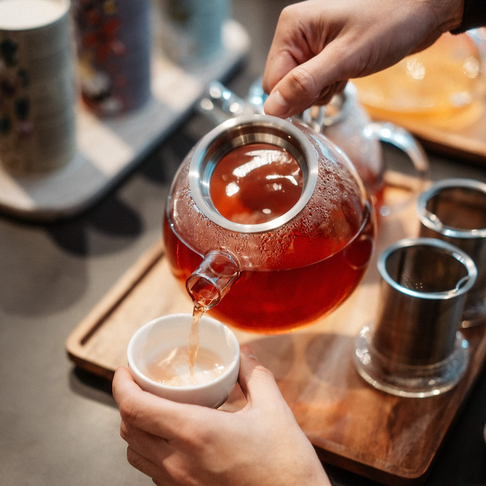 Hand gießt rötlich-braunen Tee aus Glaskanne in weiße Keramiktasse auf Holztablett mit Tee-Einsätzen.
